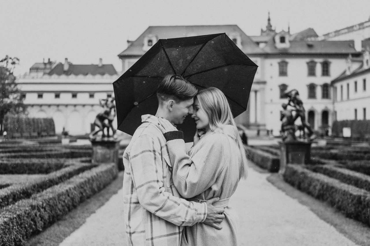 Prague couple photoshoot: a black-and-white embrace among the hedges Black-and-white wide photograph of the couple embracing under an umbrella between garden hedges during their Prague couple photoshoot.