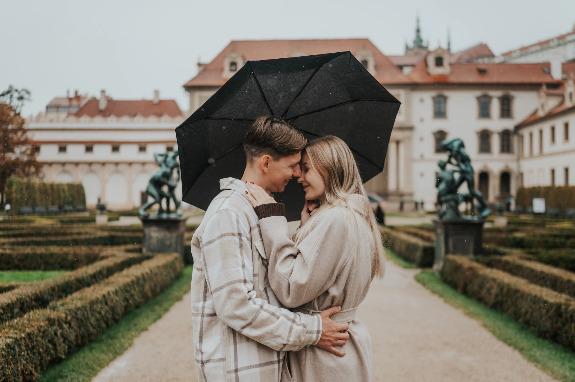 Prague couple photoshoot: an umbrella embrace with the loggia behind Wide photograph of the couple embracing under an umbrella in front of the garden loggia during their Prague couple photoshoot.