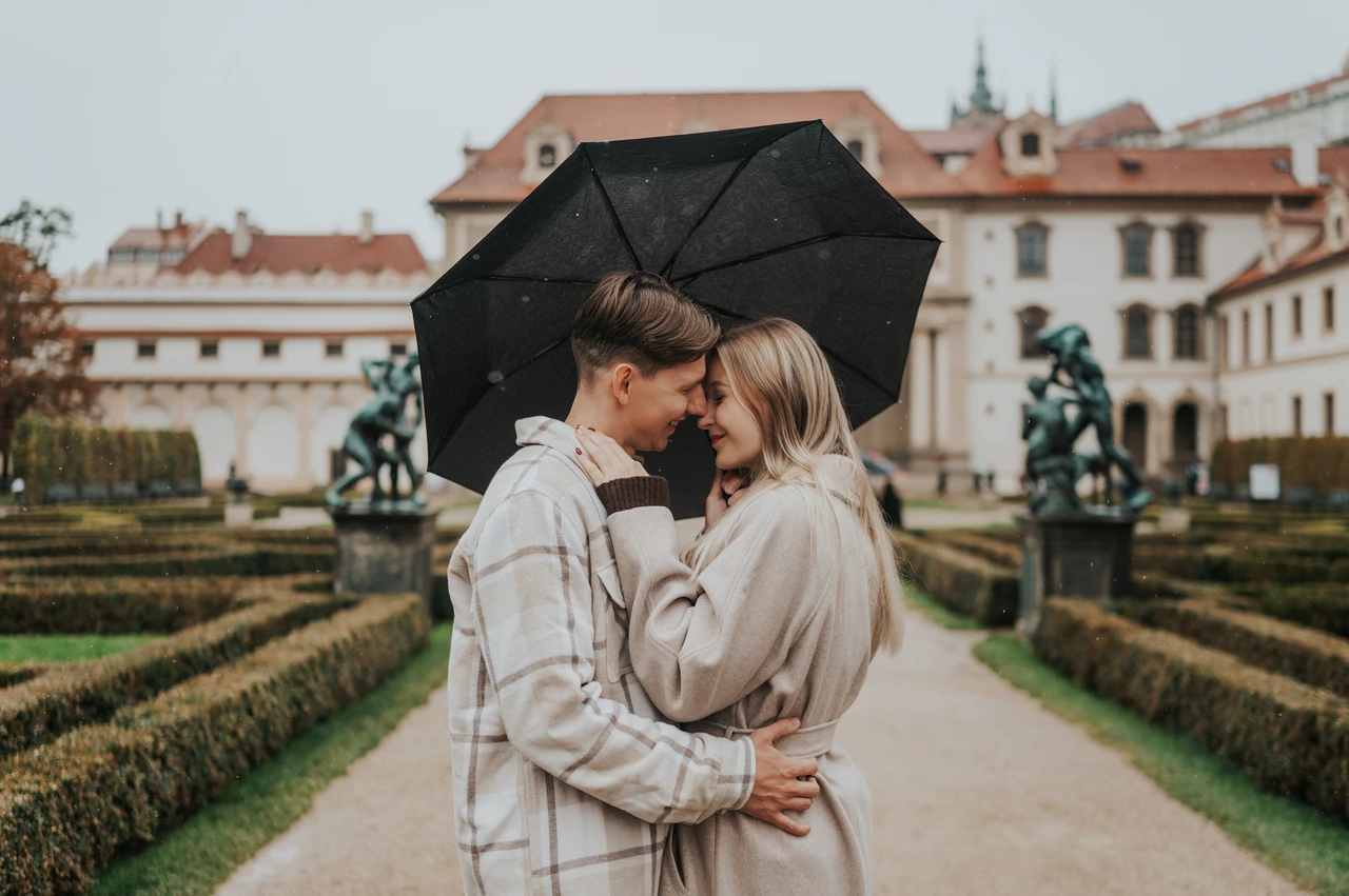 Prague couple photoshoot: an umbrella embrace with the loggia behind Wide photograph of the couple embracing under an umbrella in front of the garden loggia during their Prague couple photoshoot.