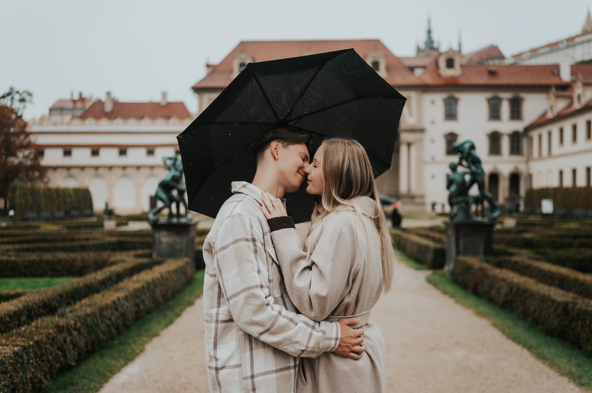 Wide photograph of the couple kissing beneath an umbrella with Wallenstein Garden's loggia behind them during their Prague couple photoshoot.