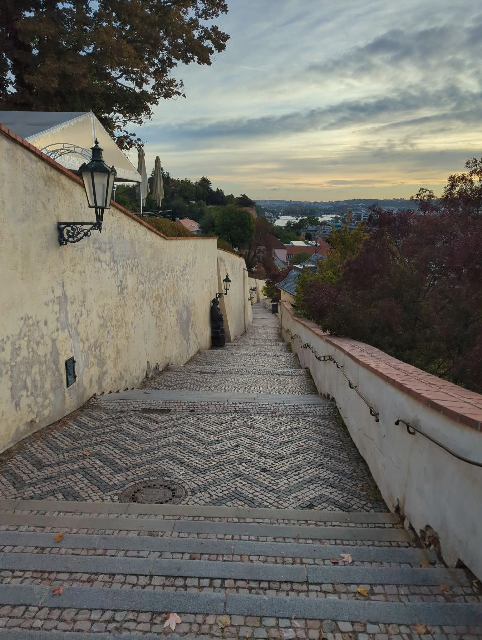 A quiet stepped lane below Prague Castle opens towards rooftops and bridges in the soft early morning.