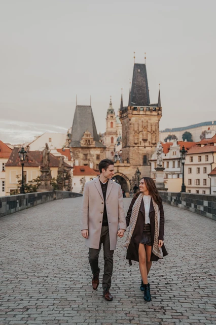 Charles Bridge engagement photoshoot in Prague Couple walking hand in hand on Charles Bridge during their Prague engagement photoshoot, with the Malá Strana towers behind them
