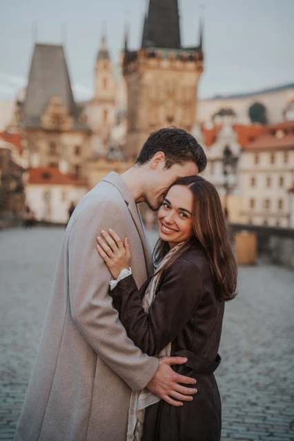 A quiet moment on Charles Bridge Woman smiling as her partner leans towards her on Charles Bridge at their engagement photoshoot in Prague