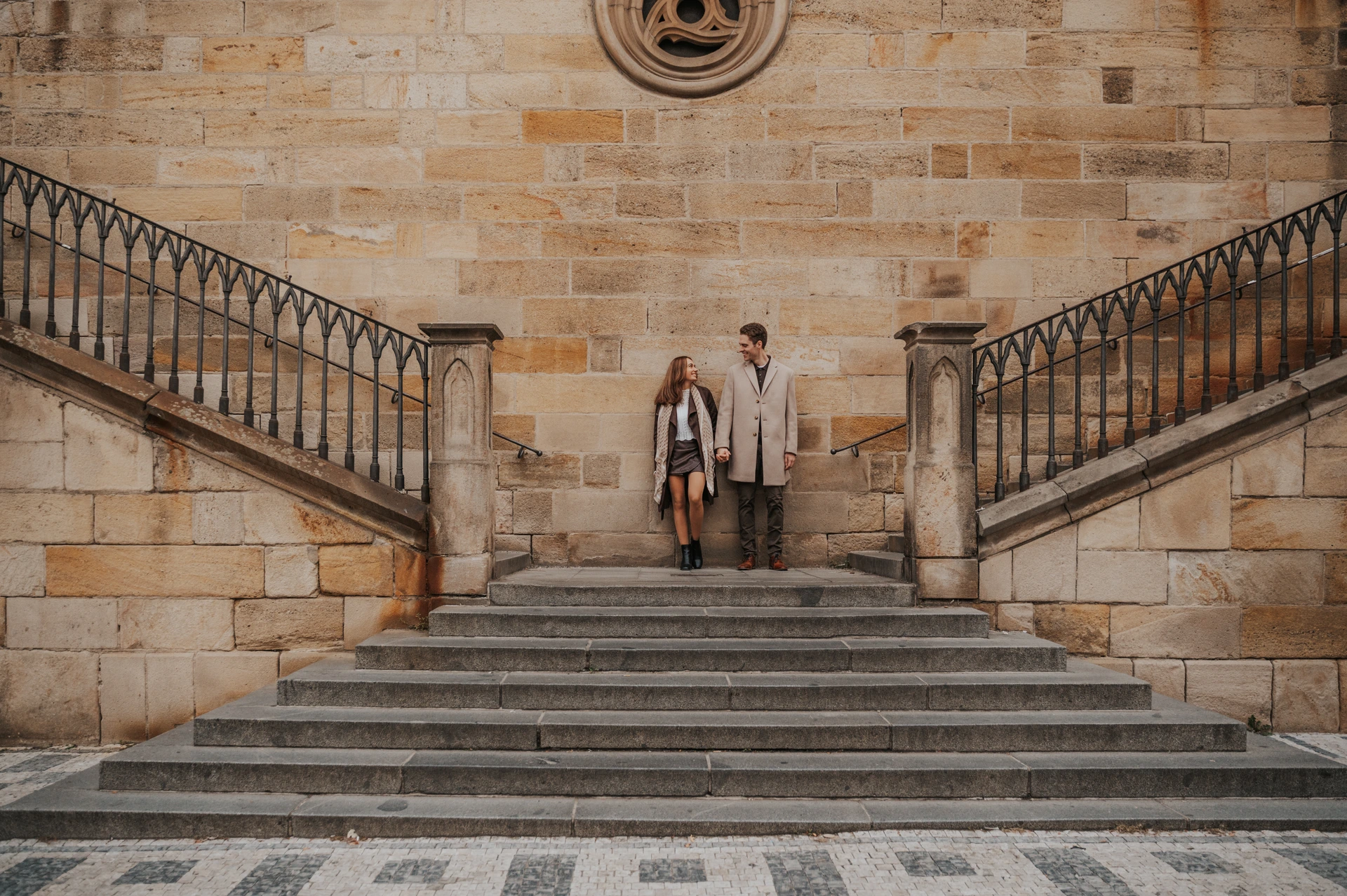 Prague engagement photoshoot by Charles Bridge Wide view of the couple on symmetrical stone stairs beside Charles Bridge during their Prague engagement photoshoot