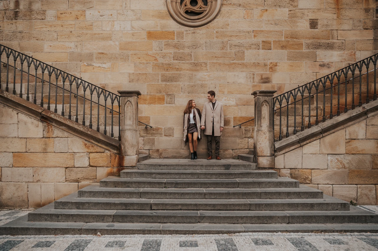 Prague engagement photoshoot by Charles Bridge Wide view of the couple on symmetrical stone stairs beside Charles Bridge during their Prague engagement photoshoot