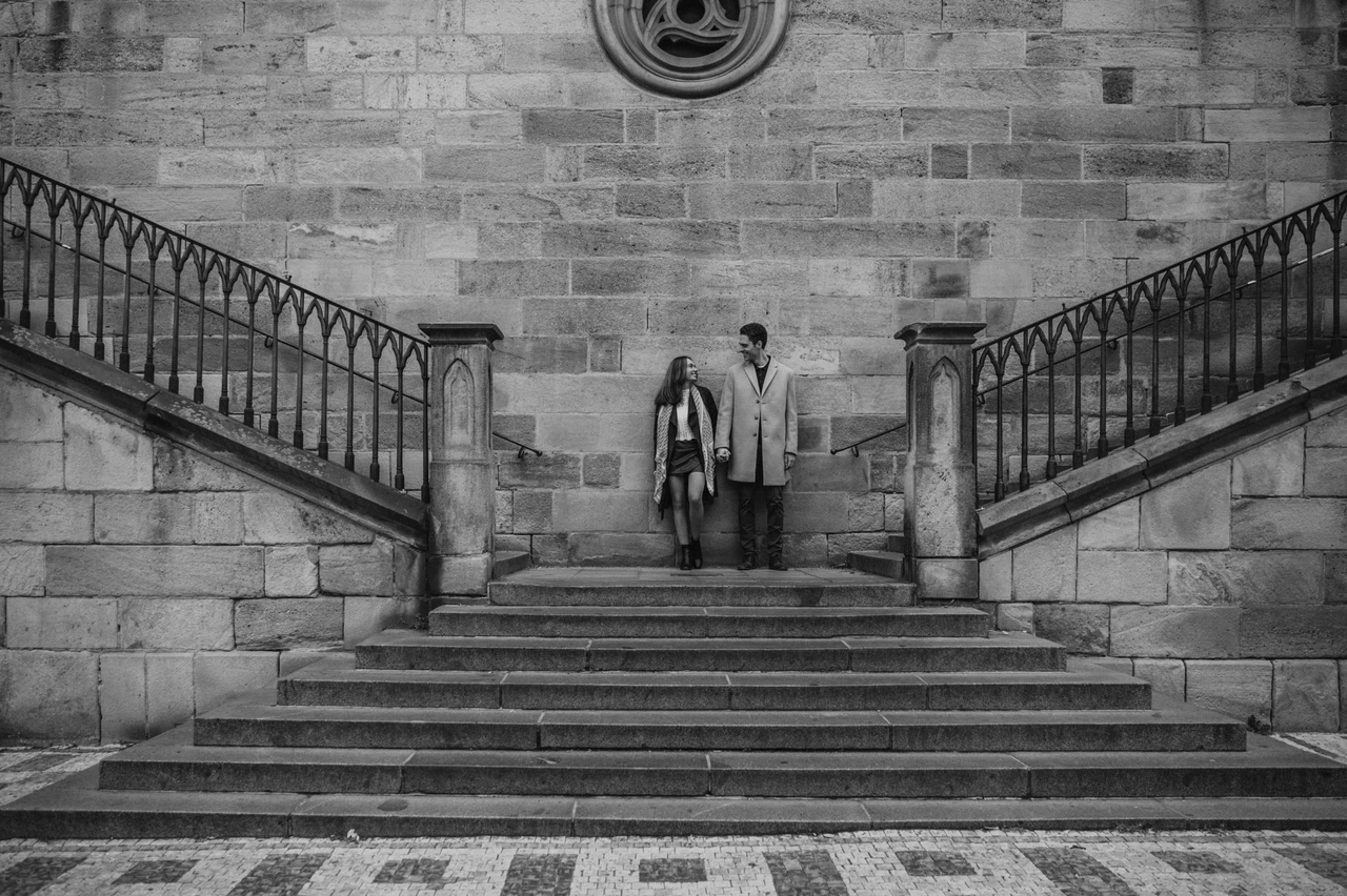 The Charles Bridge stairs in black and white Black and white wide view of the couple on the Charles Bridge stairs during an engagement photoshoot in Prague