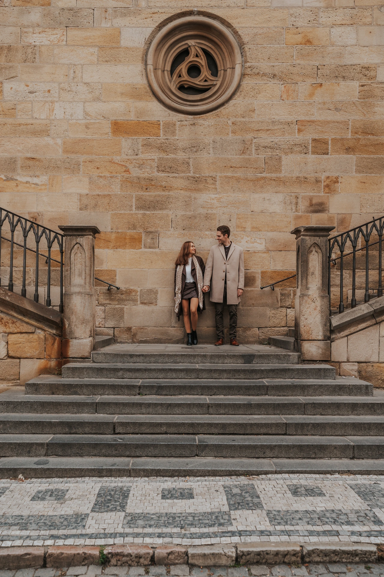 Couple standing below a round window on the Charles Bridge stairs at their Prague engagement photoshoot