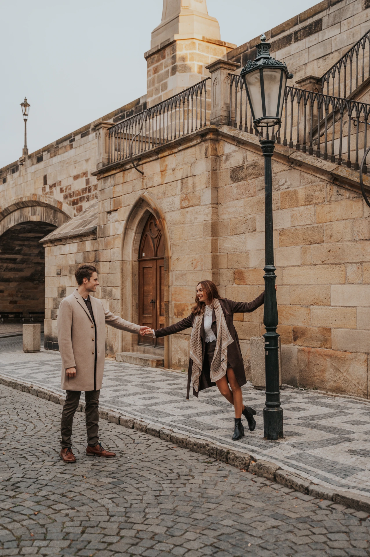Couple smiling beside stone walls and lamps below Charles Bridge at their Prague engagement photoshoot