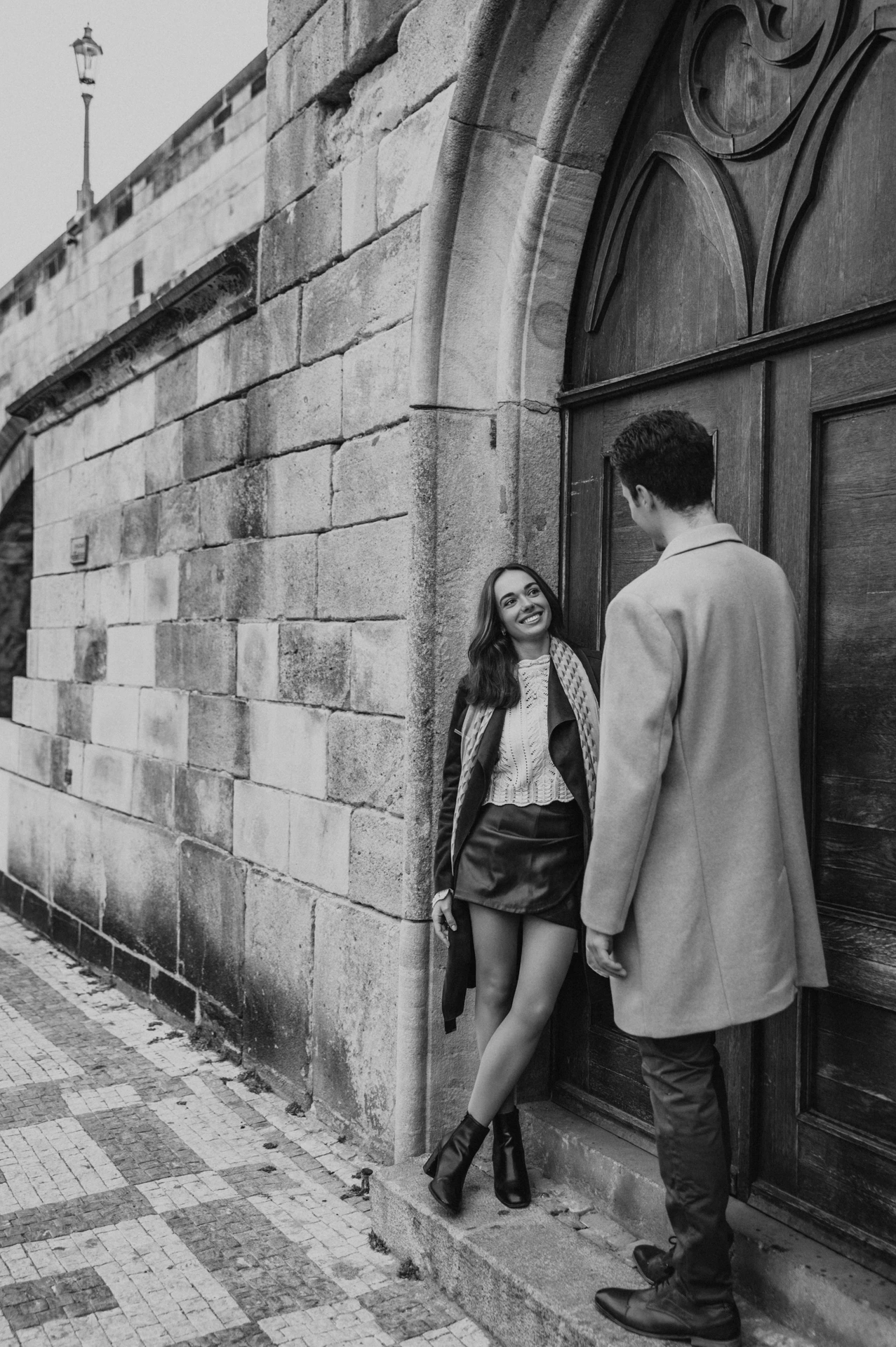 Black and white portrait of the couple at an arched wooden door during their Prague engagement photoshoot