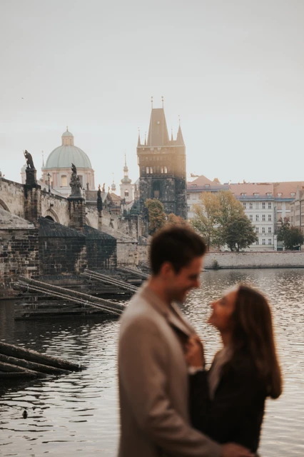 Charles Bridge beyond a riverside portrait Soft-focus foreground portrait of the couple with Charles Bridge across the Vltava during their Prague engagement photoshoot