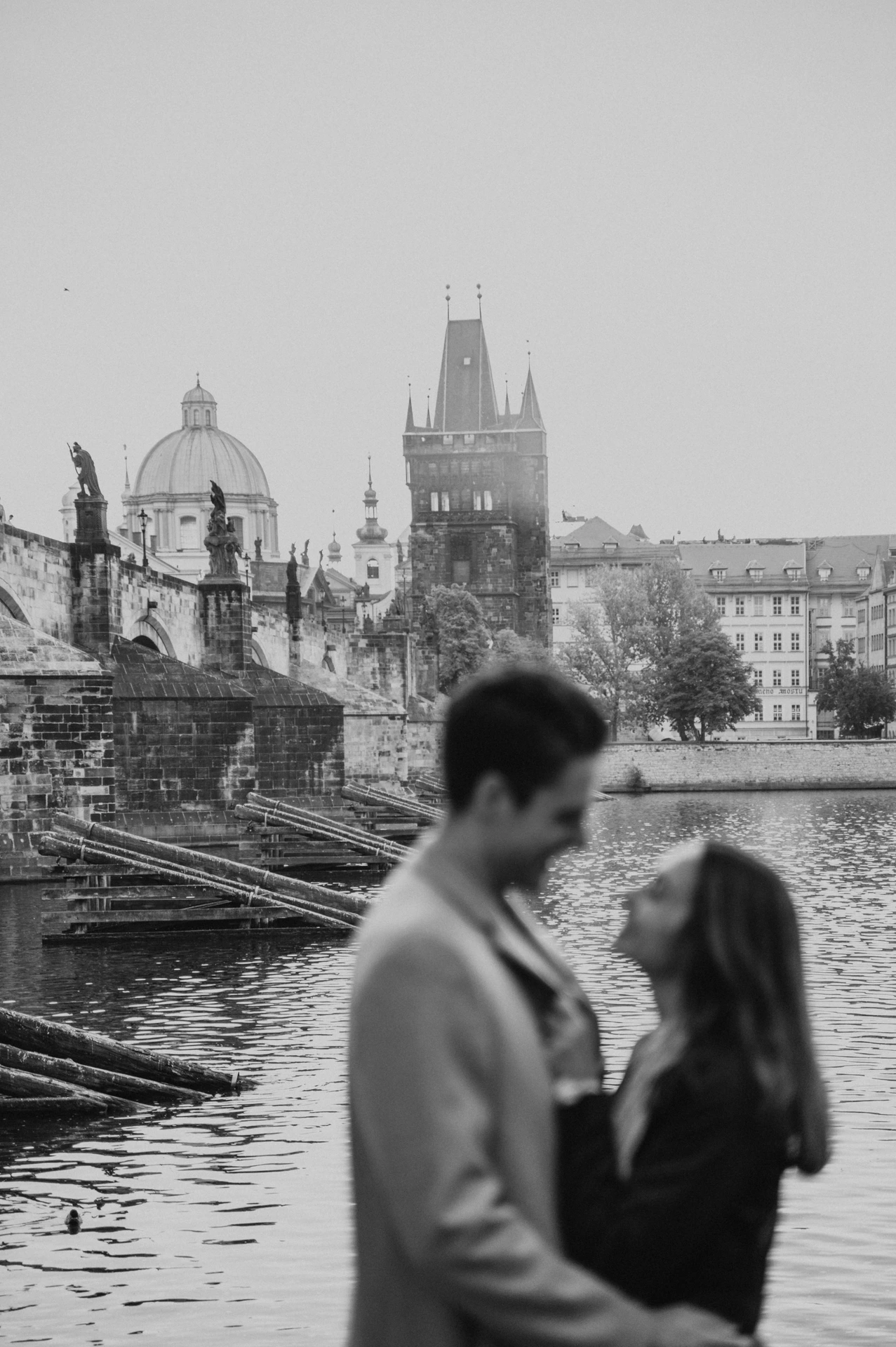 Black and white portrait of the couple beside the Vltava during their Prague engagement photoshoot, with Charles Bridge behind them