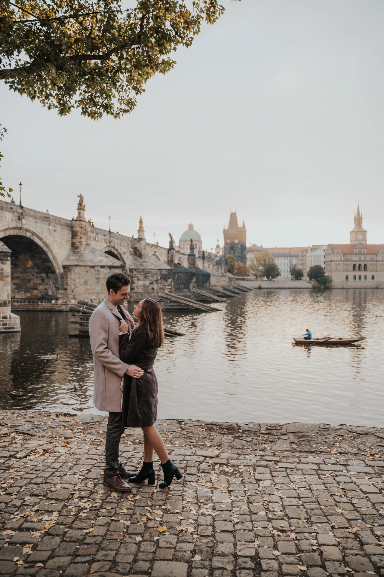 Couple embracing on the cobbled Kampa riverbank with Charles Bridge behind them during their Prague engagement photoshoot