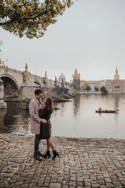 Engagement photoshoot on the Kampa riverbank Couple embracing on the cobbled Kampa riverbank with Charles Bridge behind them during their Prague engagement photoshoot