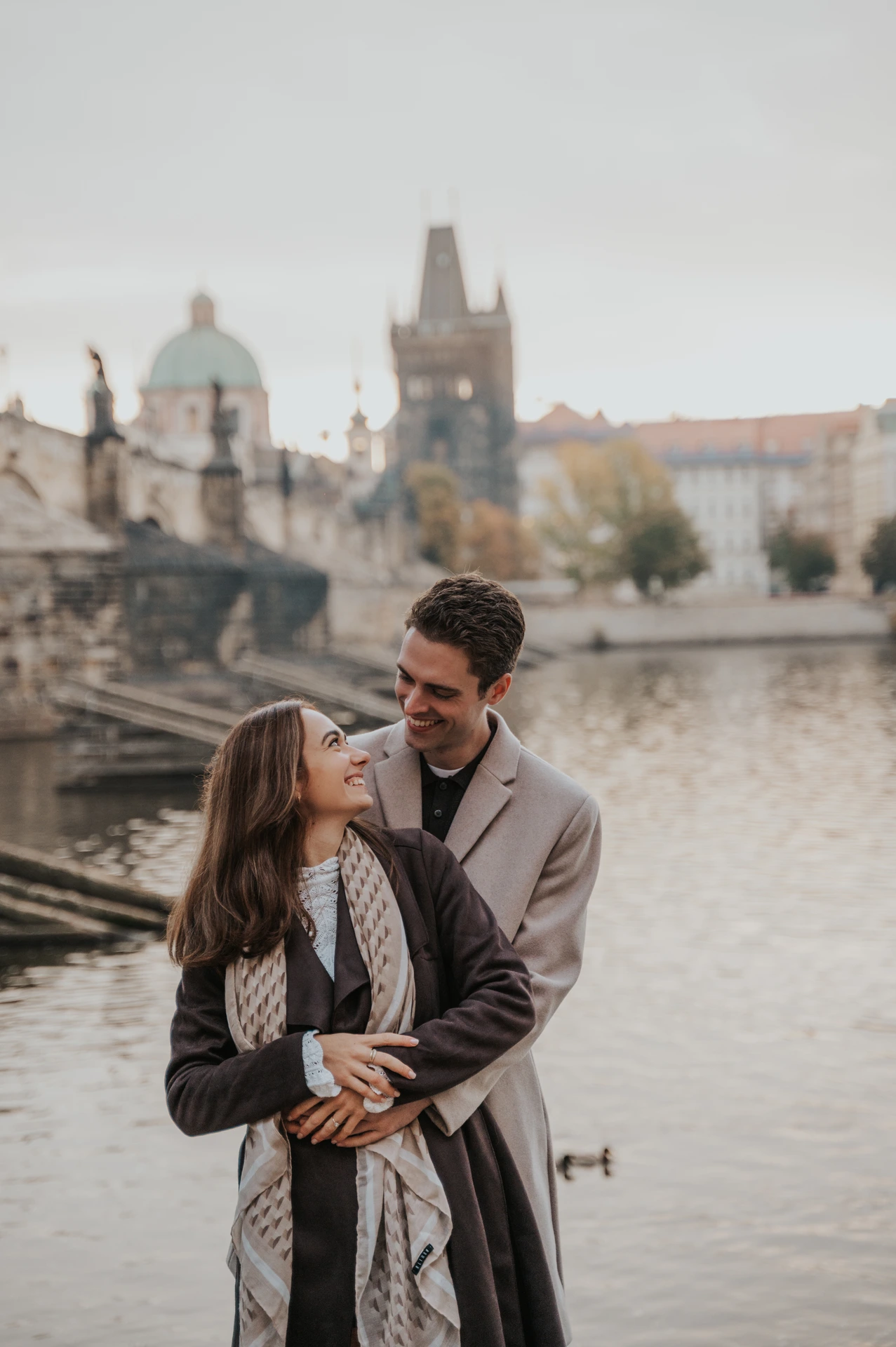Couple hugging beside the Vltava with Charles Bridge in the background at their Prague engagement photoshoot
