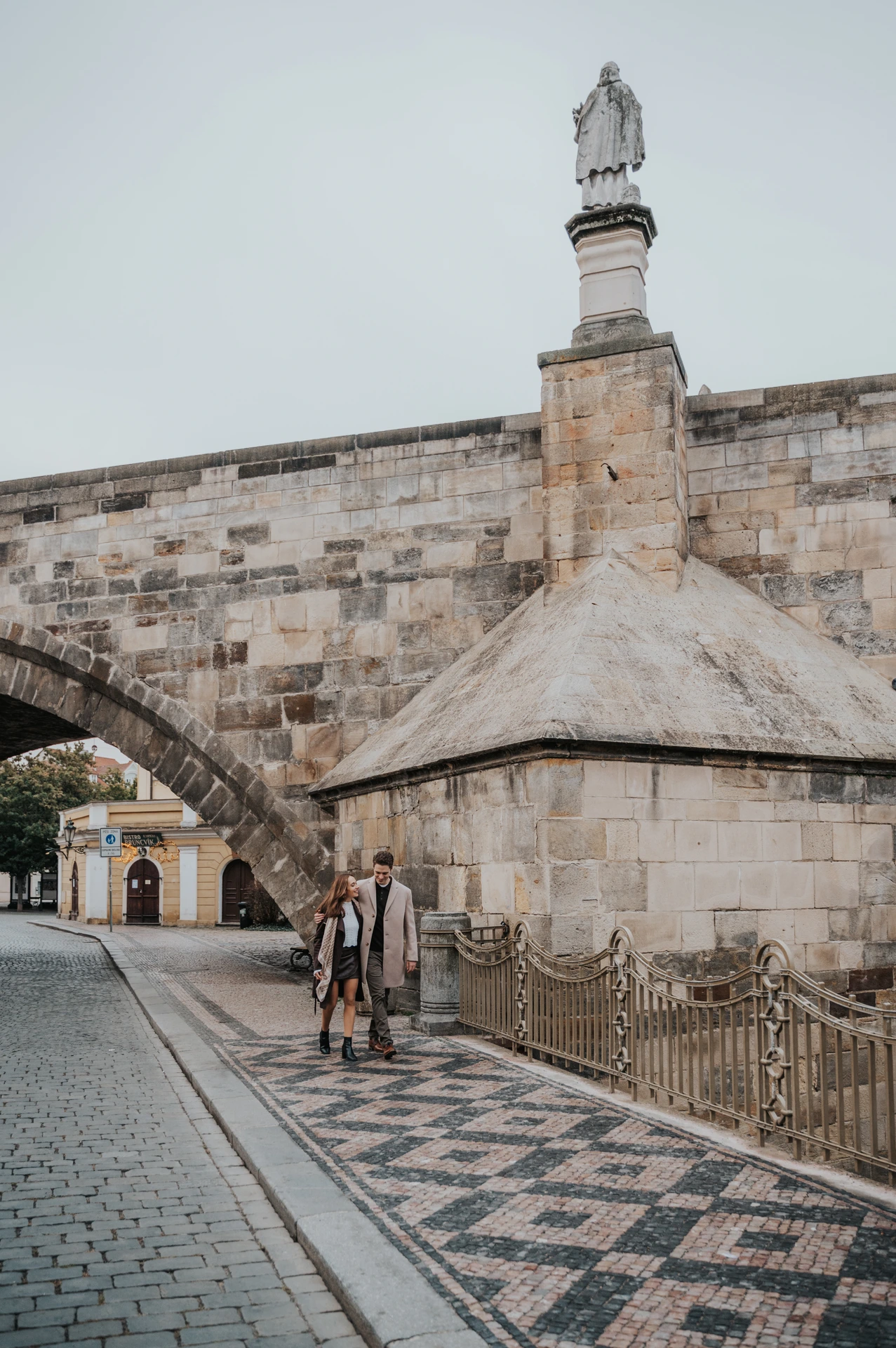 Couple walking below a stone arch of Charles Bridge during their Prague engagement photoshoot