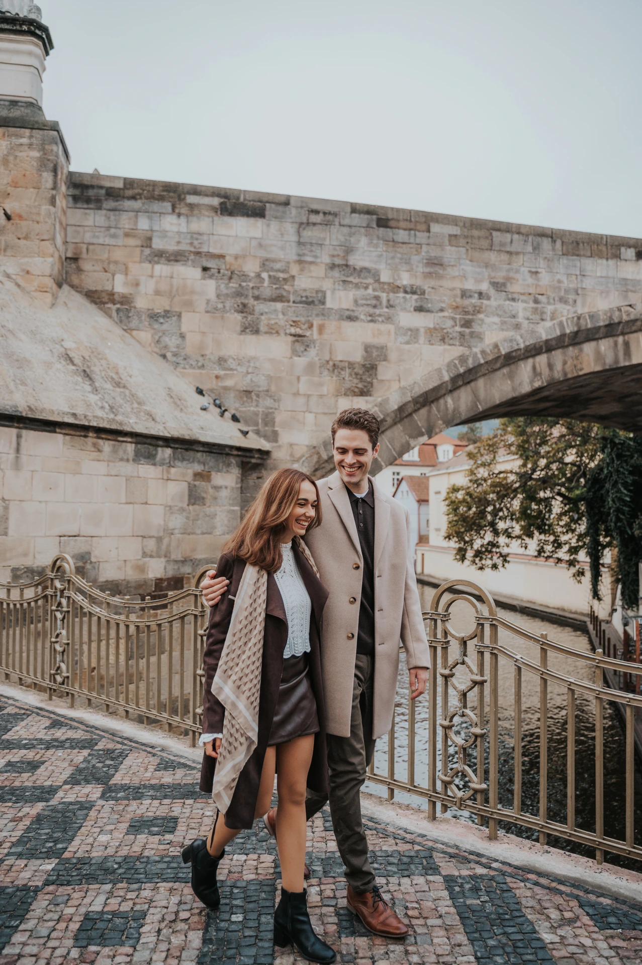 Couple smiling side by side below the Charles Bridge arches during their Prague engagement photoshoot