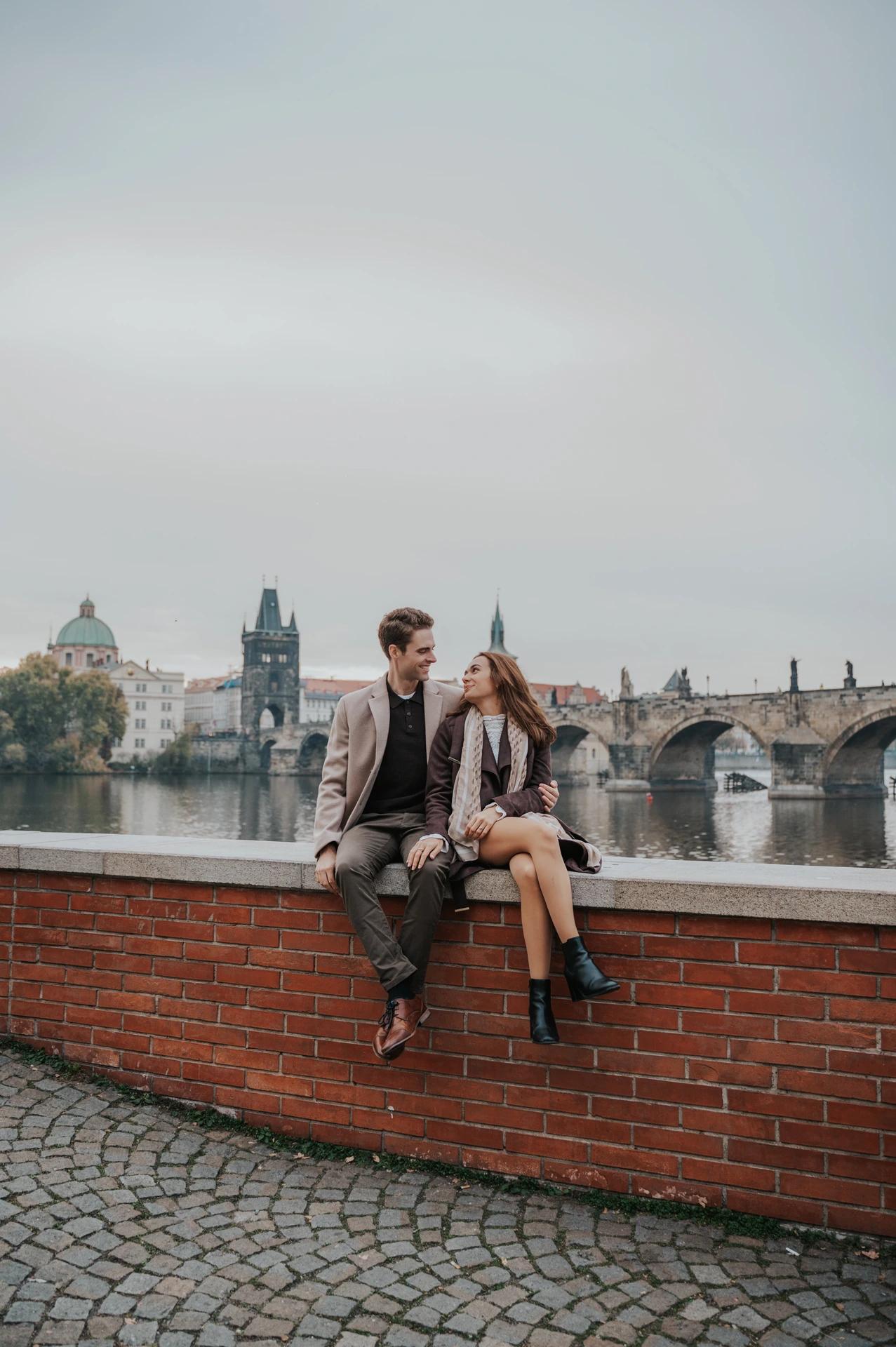 Couple sitting on a riverside brick wall with Charles Bridge behind them during their Prague engagement photoshoot
