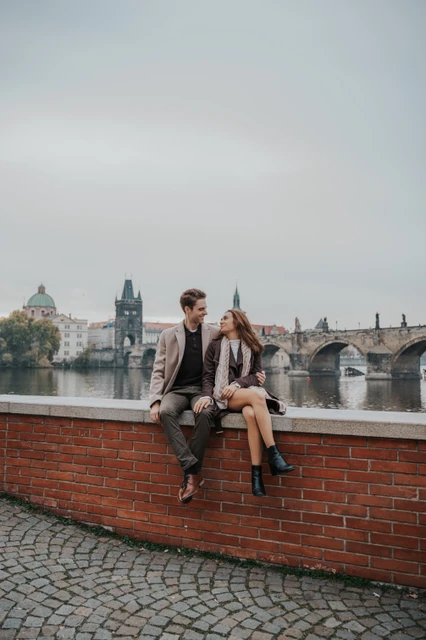 An engagement portrait beside the Vltava Couple sitting on a riverside brick wall with Charles Bridge behind them during their Prague engagement photoshoot