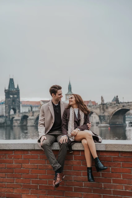 Riverside smiles with Charles Bridge behind Couple smiling at each other while sitting on a wall by the Vltava at their Prague engagement photoshoot