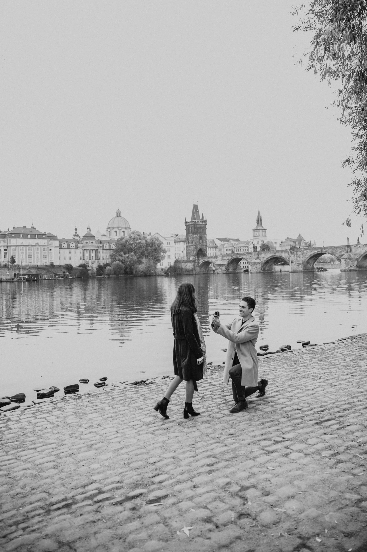 Black and white photograph of the man on one knee during the couple's proposal photoshoot on the Kampa riverbank in Prague