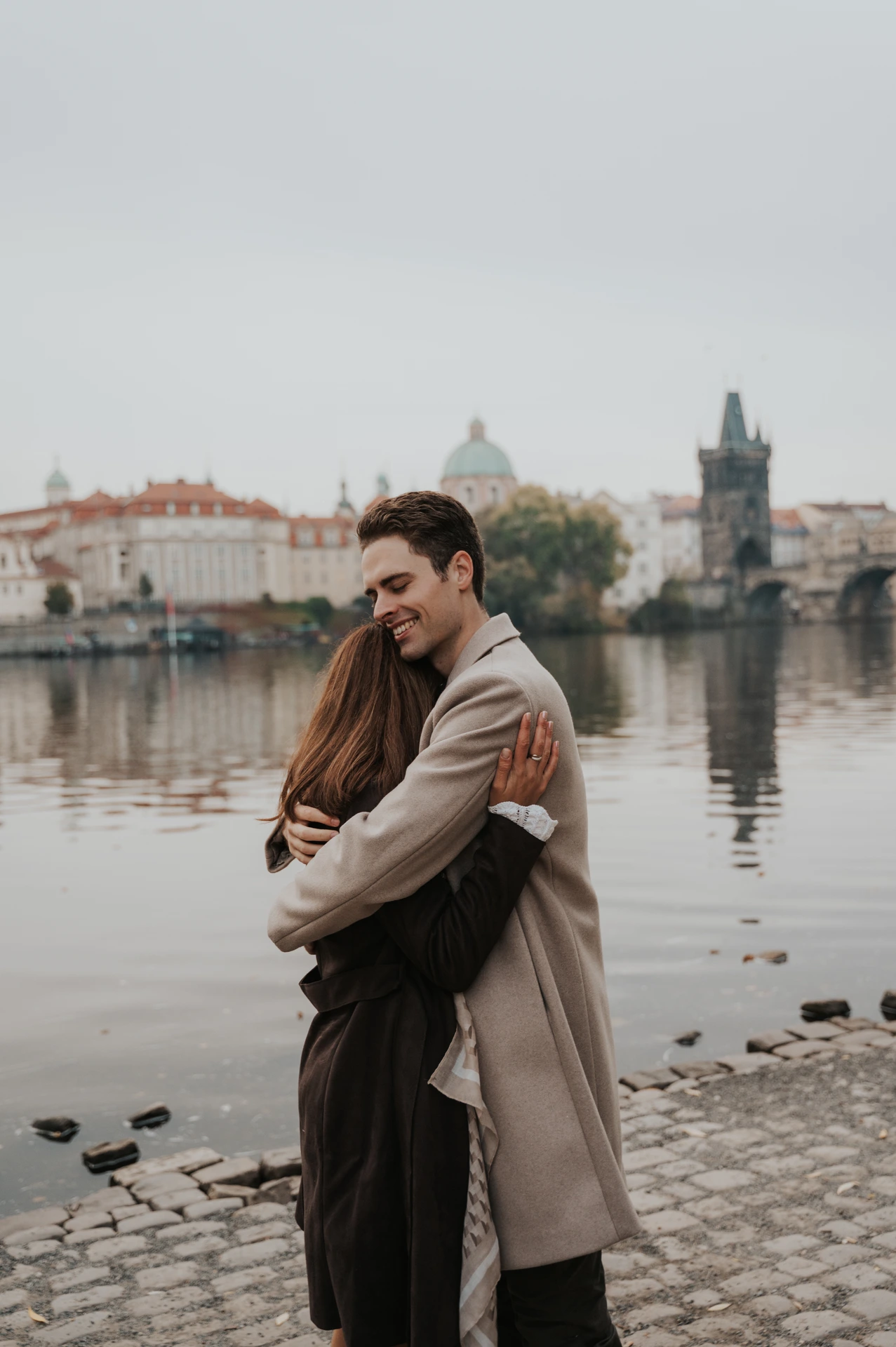 Newly engaged couple embracing beside the Vltava during their Prague engagement photoshoot