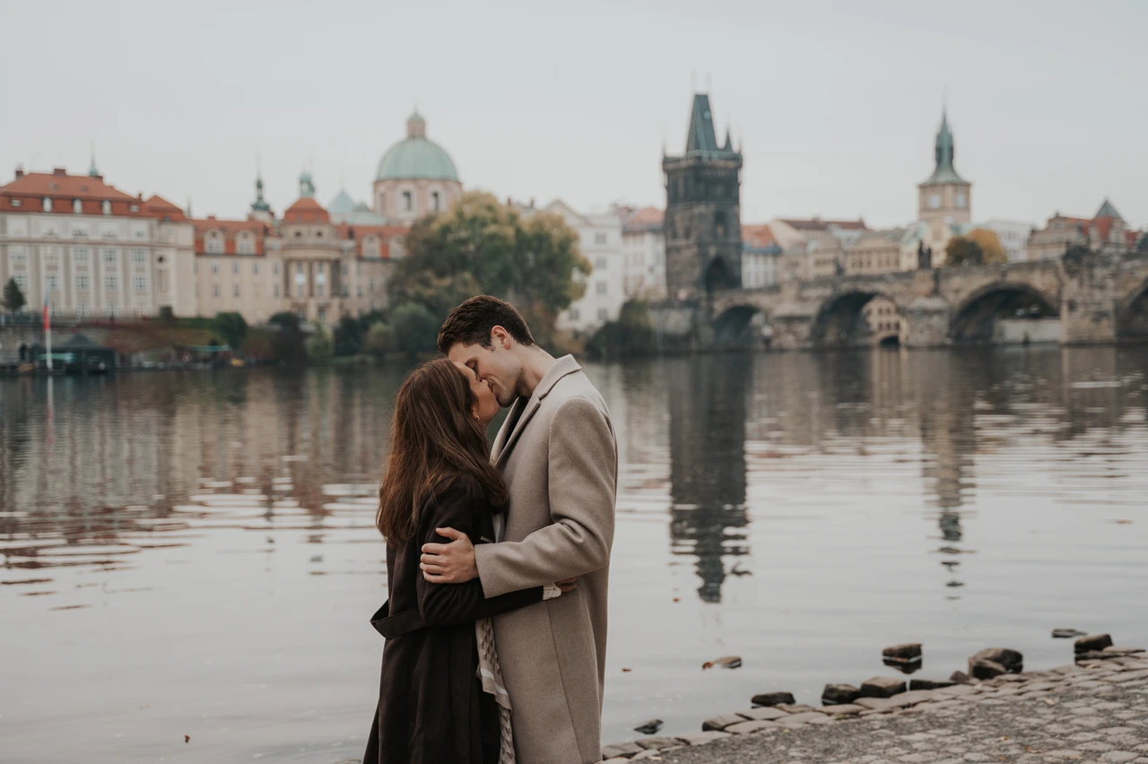 A riverside kiss at their Prague engagement photoshoot Wide view of the newly engaged couple kissing beside the Vltava, with Charles Bridge behind them, during their Prague engagement photoshoot