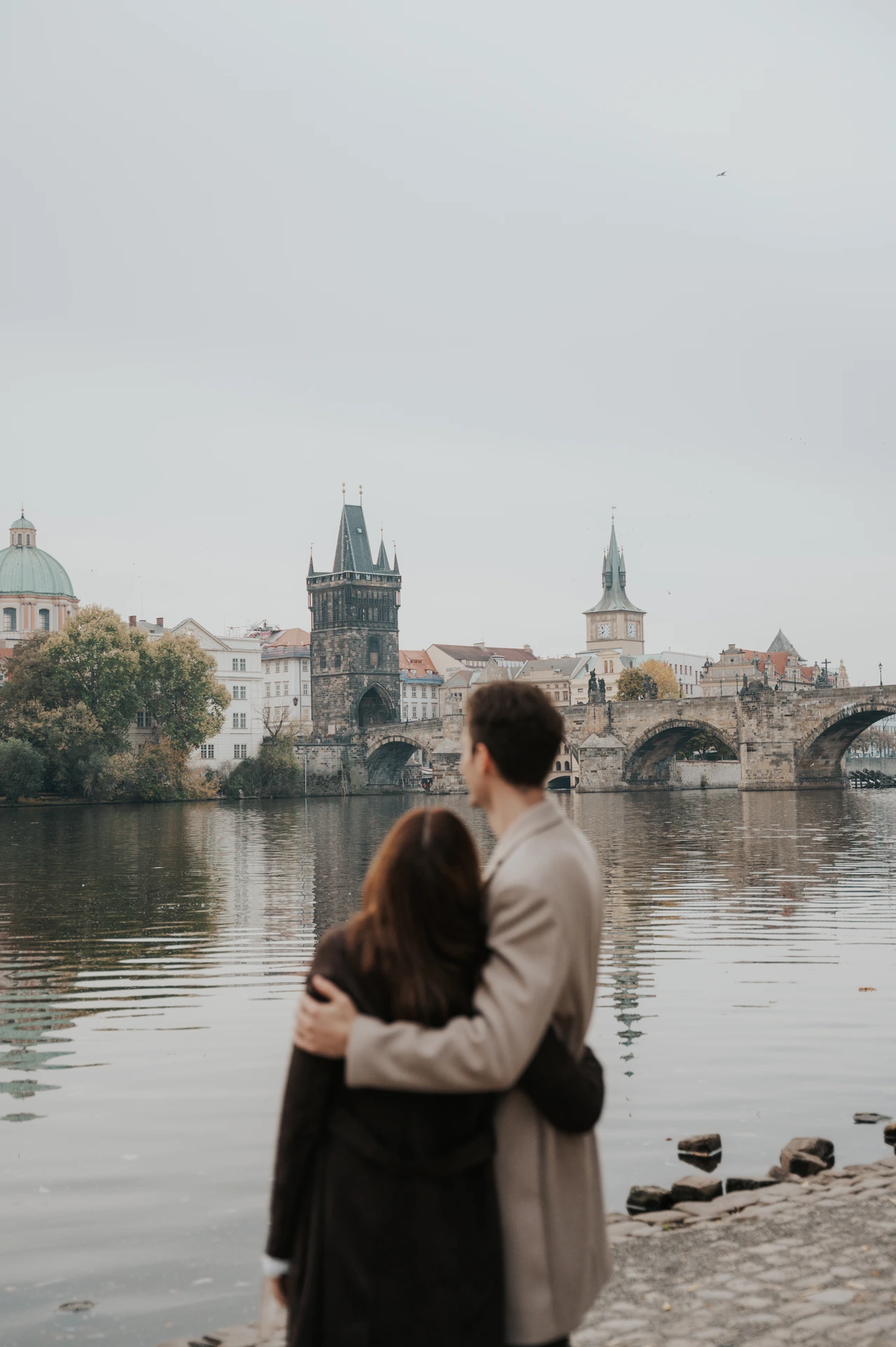 Couple embracing and looking towards Charles Bridge across the Vltava during their Prague engagement photoshoot