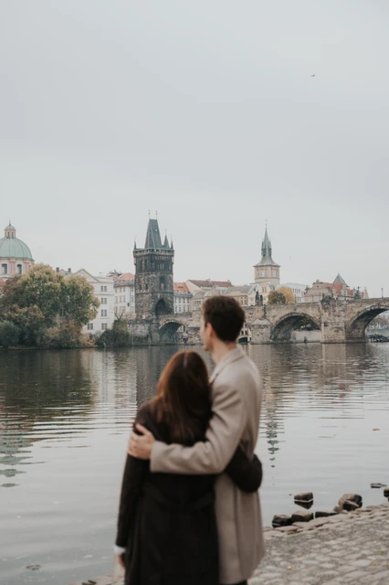 Looking towards Charles Bridge together Couple embracing and looking towards Charles Bridge across the Vltava during their Prague engagement photoshoot