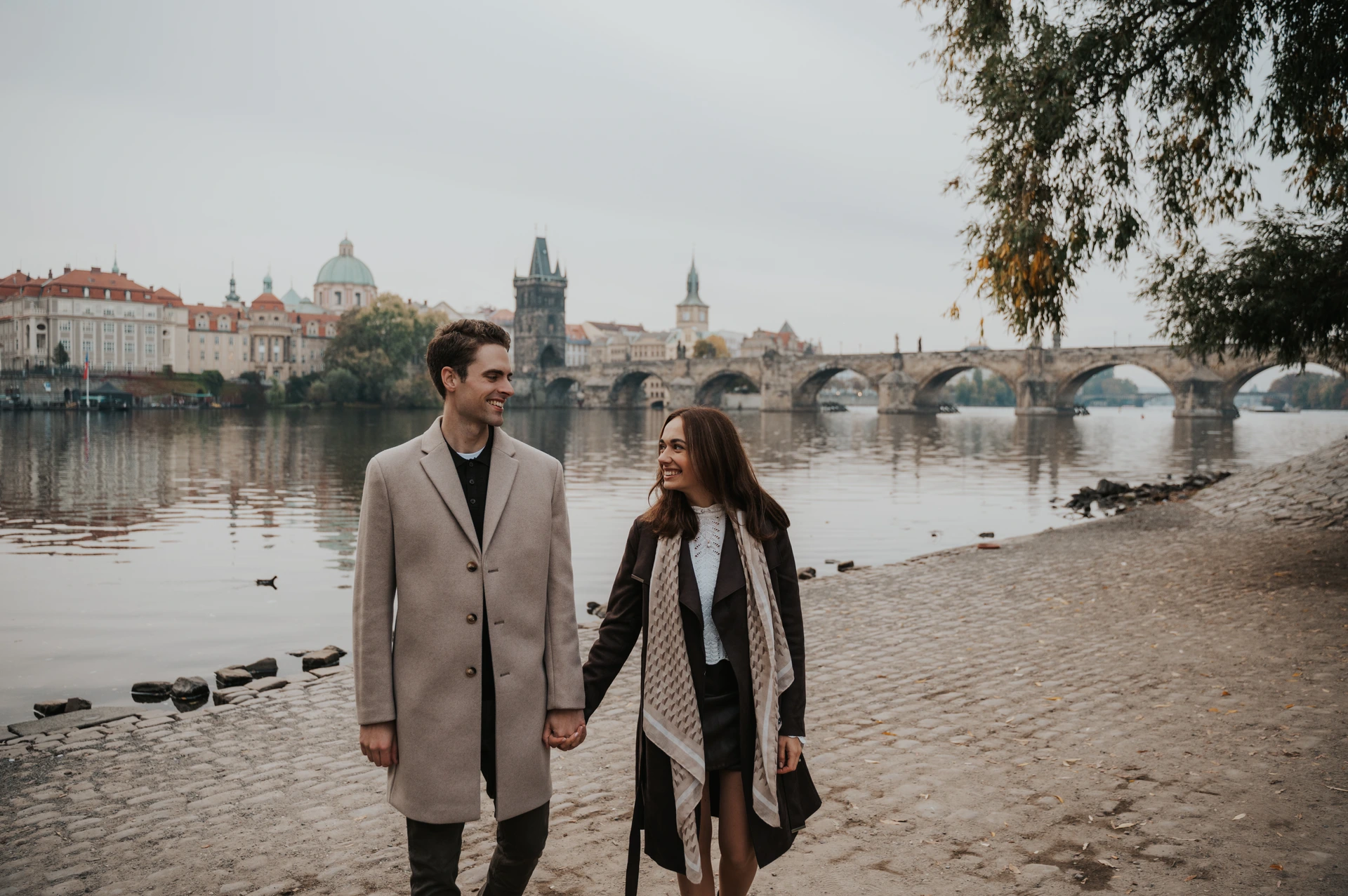 Prague engagement photoshoot along the Kampa riverbank Wide photograph of the couple walking hand in hand along the Kampa riverbank during their Prague engagement photoshoot