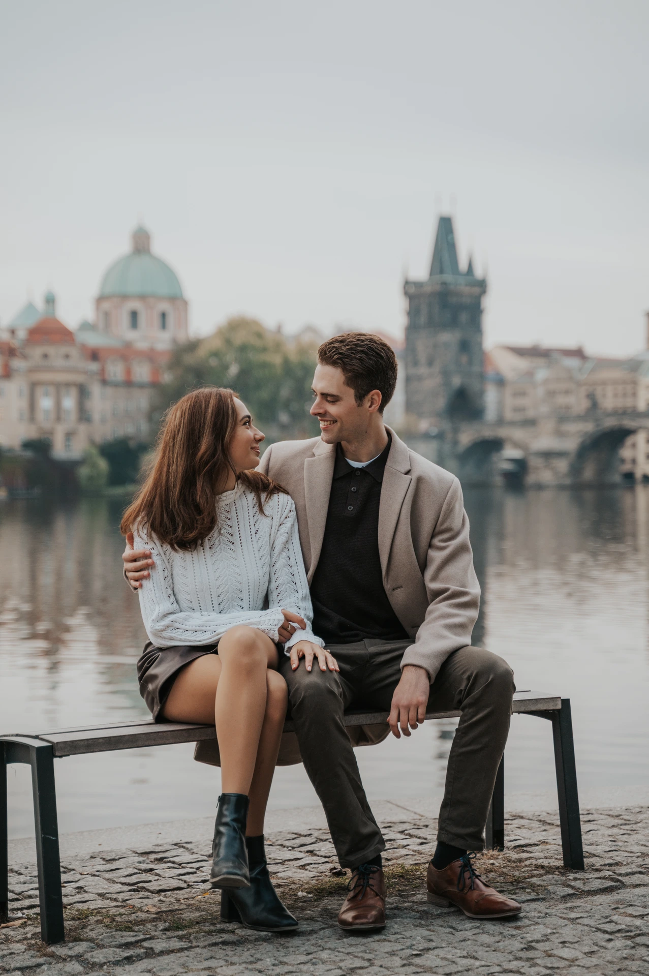Couple sitting on a riverside bench and smiling at each other during their Prague engagement photoshoot