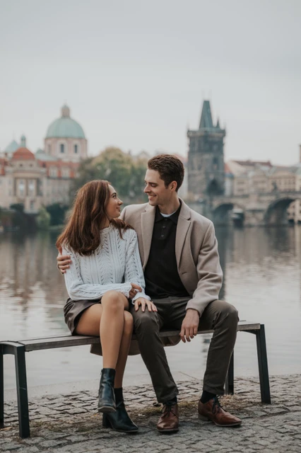 A riverside bench and a shared smile Couple sitting on a riverside bench and smiling at each other during their Prague engagement photoshoot