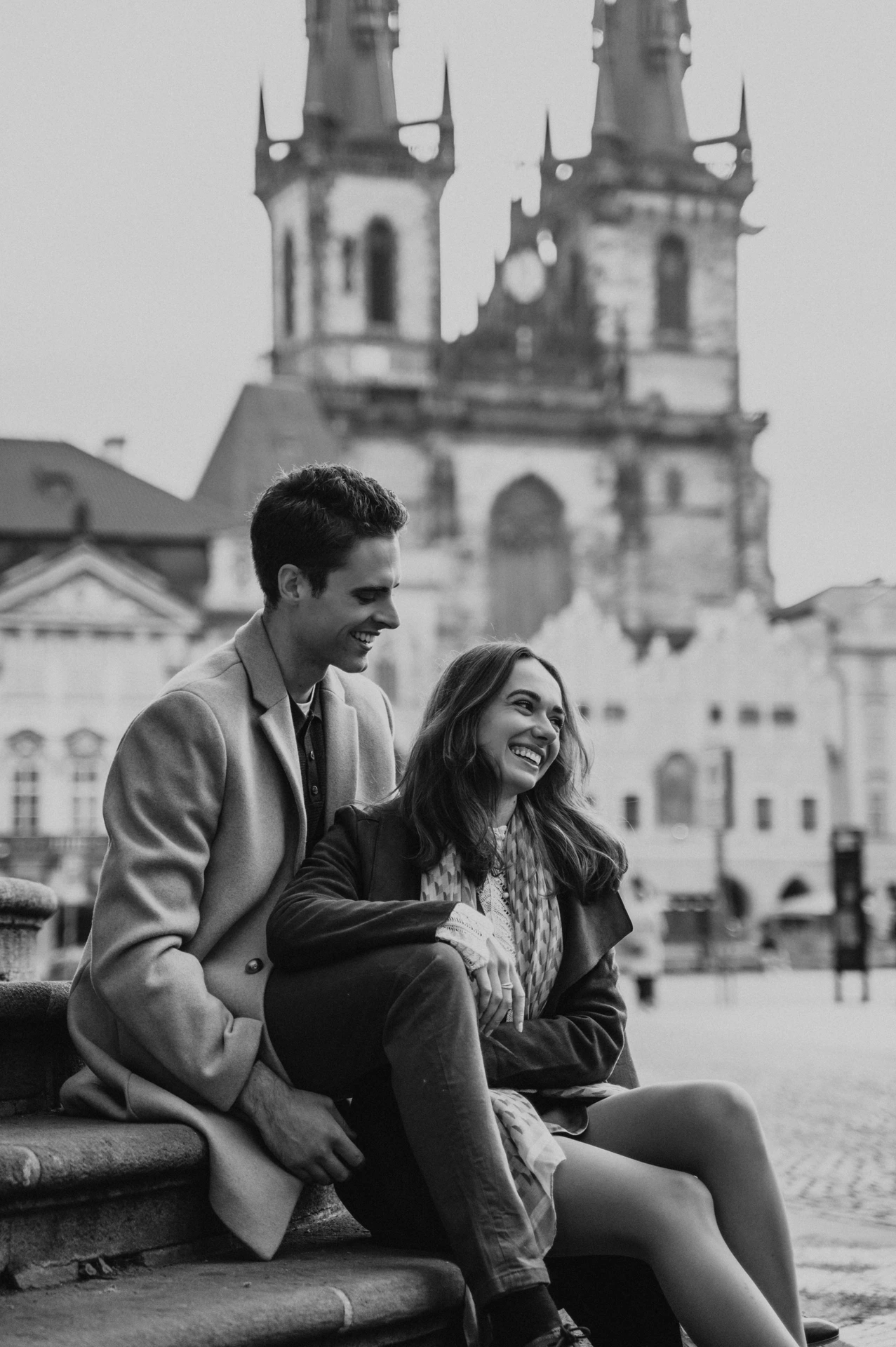 Black and white portrait of the couple laughing on steps in Old Town Square during their Prague engagement photoshoot