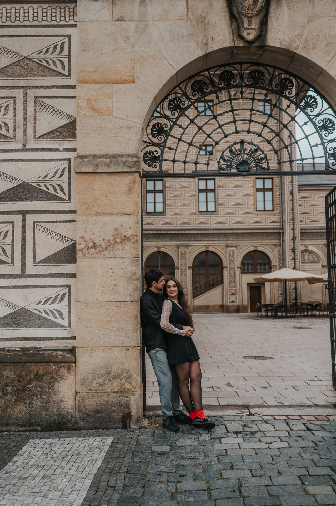 Couple embracing beside the stone arch of an iron gateway during their Prague engagement photoshoot.