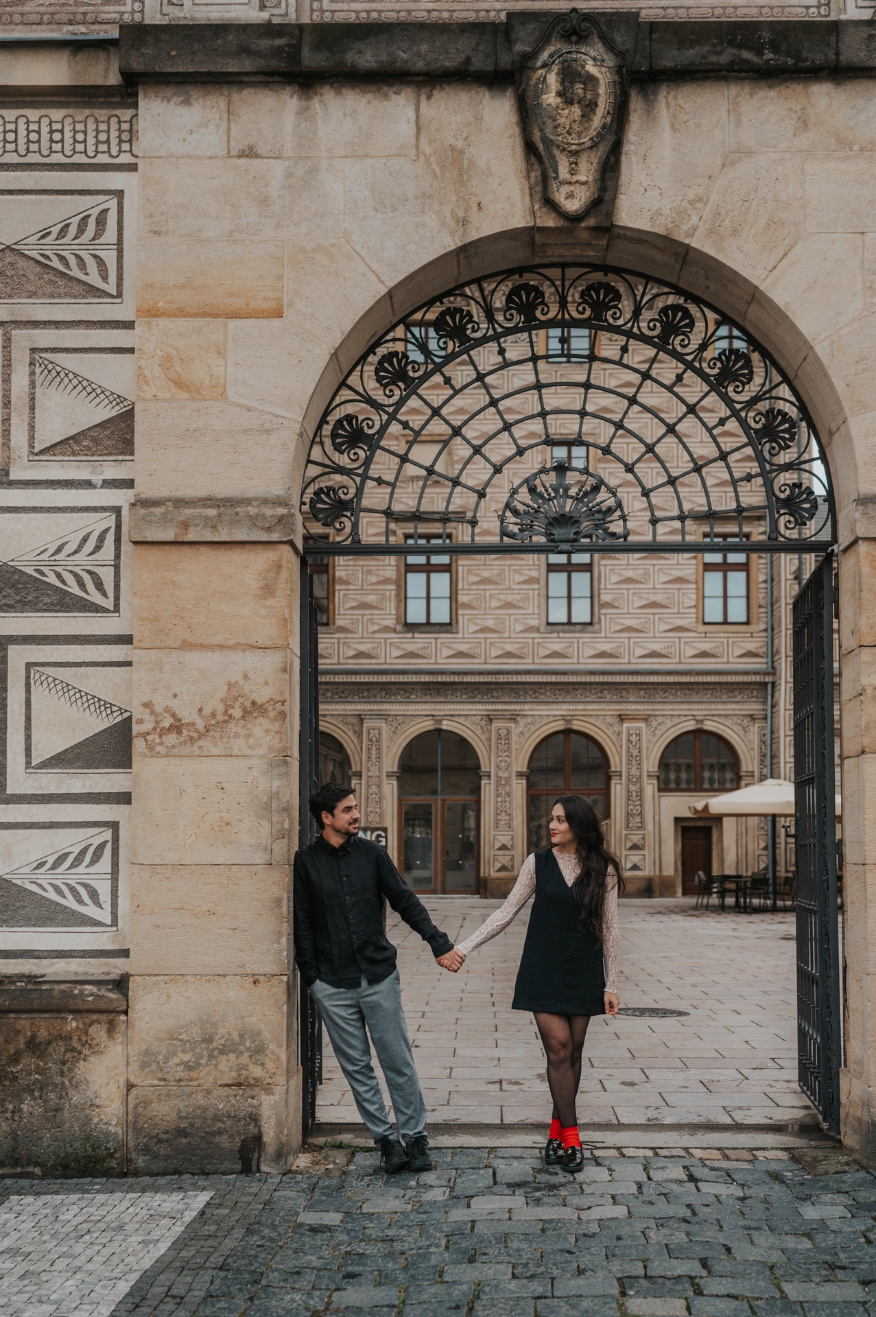 Couple holding hands across an ornate arched gateway near Prague Castle during their engagement photoshoot.