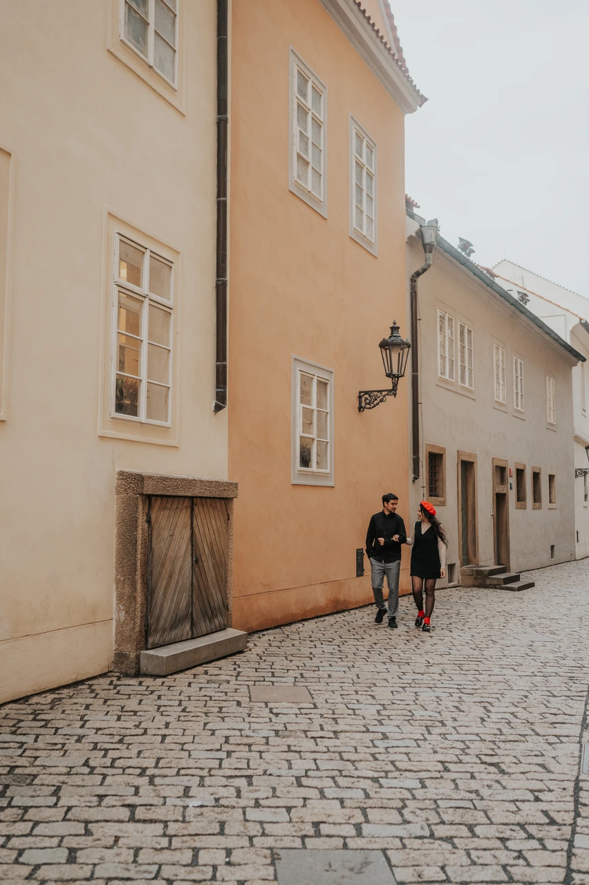 Prague engagement photoshoot on a colourful cobbled castle lane Couple walking hand in hand along a cobbled lane beside warm-coloured castle buildings during their Prague engagement photoshoot.