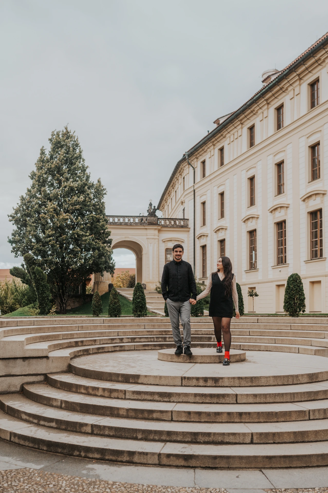 Couple standing together on broad curved steps in a castle courtyard during their Prague engagement photoshoot.