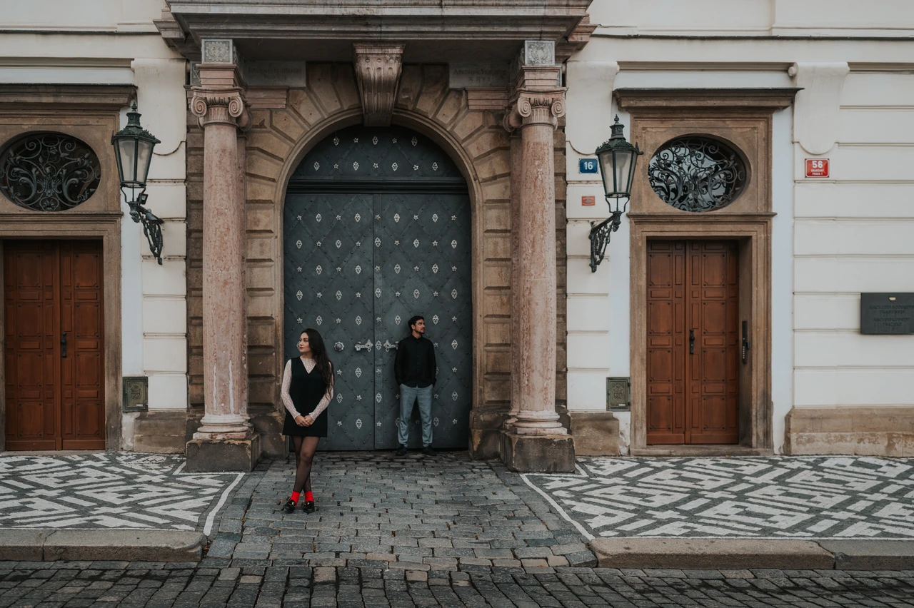 Wide Prague engagement photoshoot by a carved stone doorway Wide photograph of the couple standing by a studded door beneath a balcony in the castle district during their engagement photoshoot in Prague.