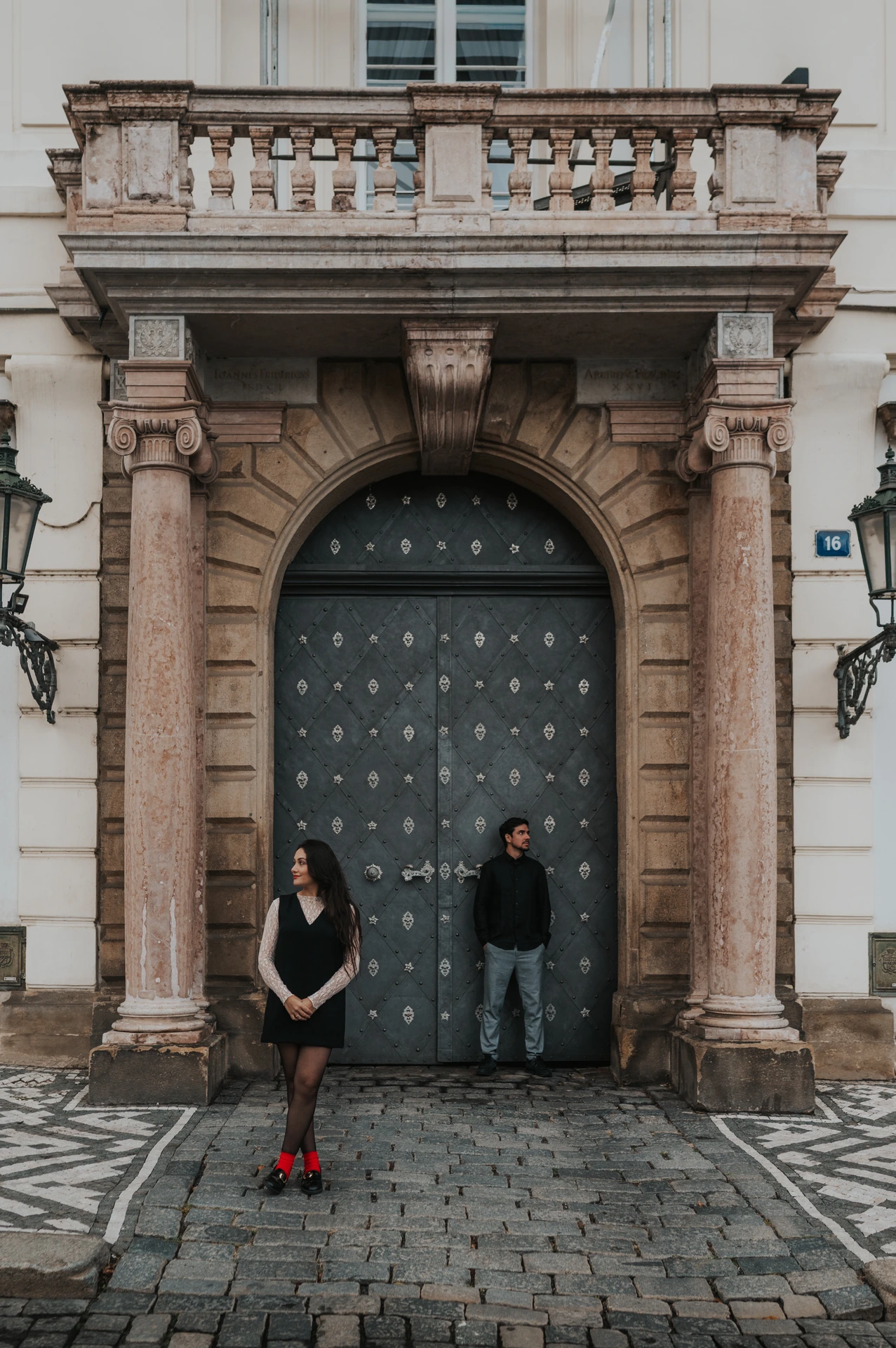 Couple standing apart in front of a tall studded door and carved stone doorway during their Prague engagement photoshoot.