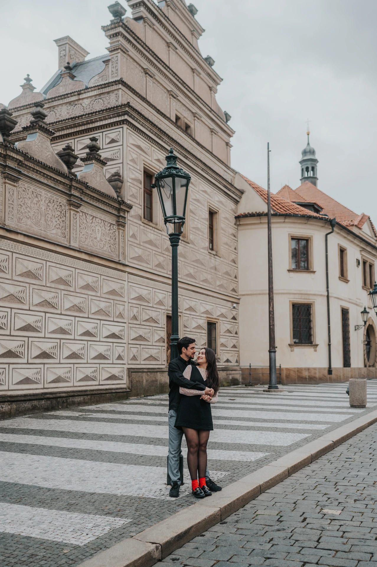 Couple embracing beside an old street lamp with castle-area architecture behind them during their engagement photoshoot in Prague.
