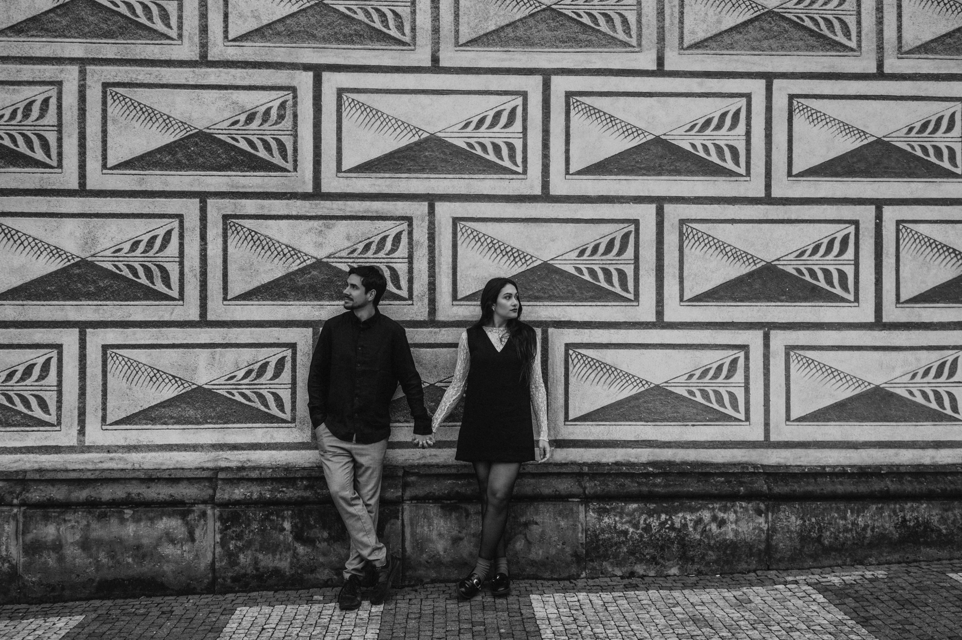 Black-and-white landscape portrait of the couple holding hands by a patterned castle-area facade during their Prague engagement photoshoot.
