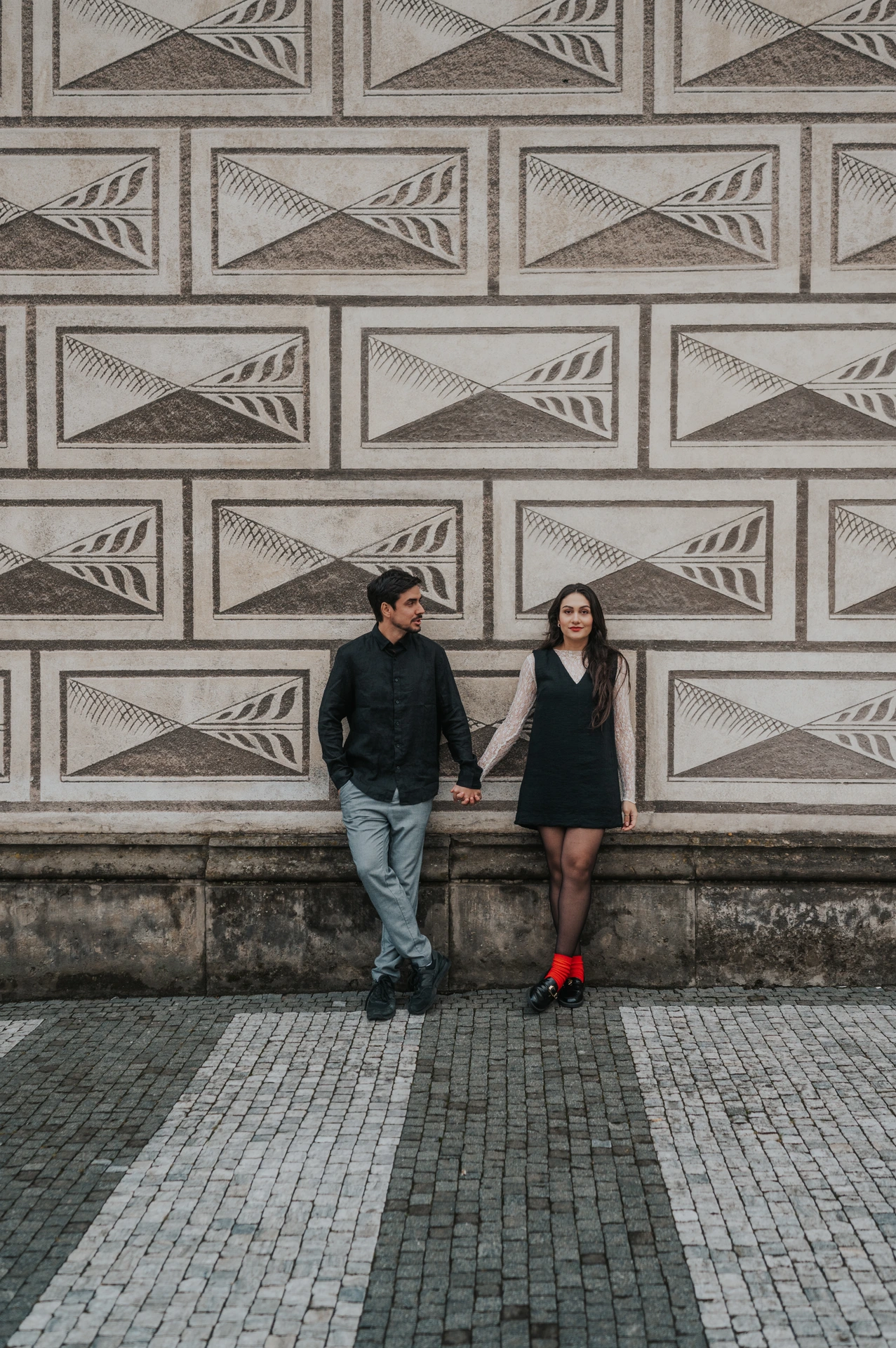 Full-length portrait of the couple looking towards each other beside a patterned wall, with her red boots visible, during their Prague engagement photoshoot.