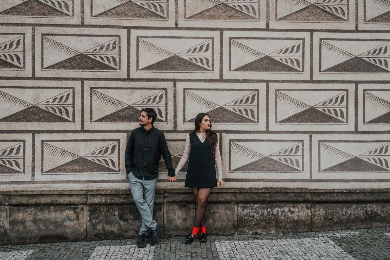 Wide Prague engagement photoshoot with a patterned stone backdrop Wide photograph of the couple holding hands against a patterned facade near Prague Castle during their engagement photoshoot.