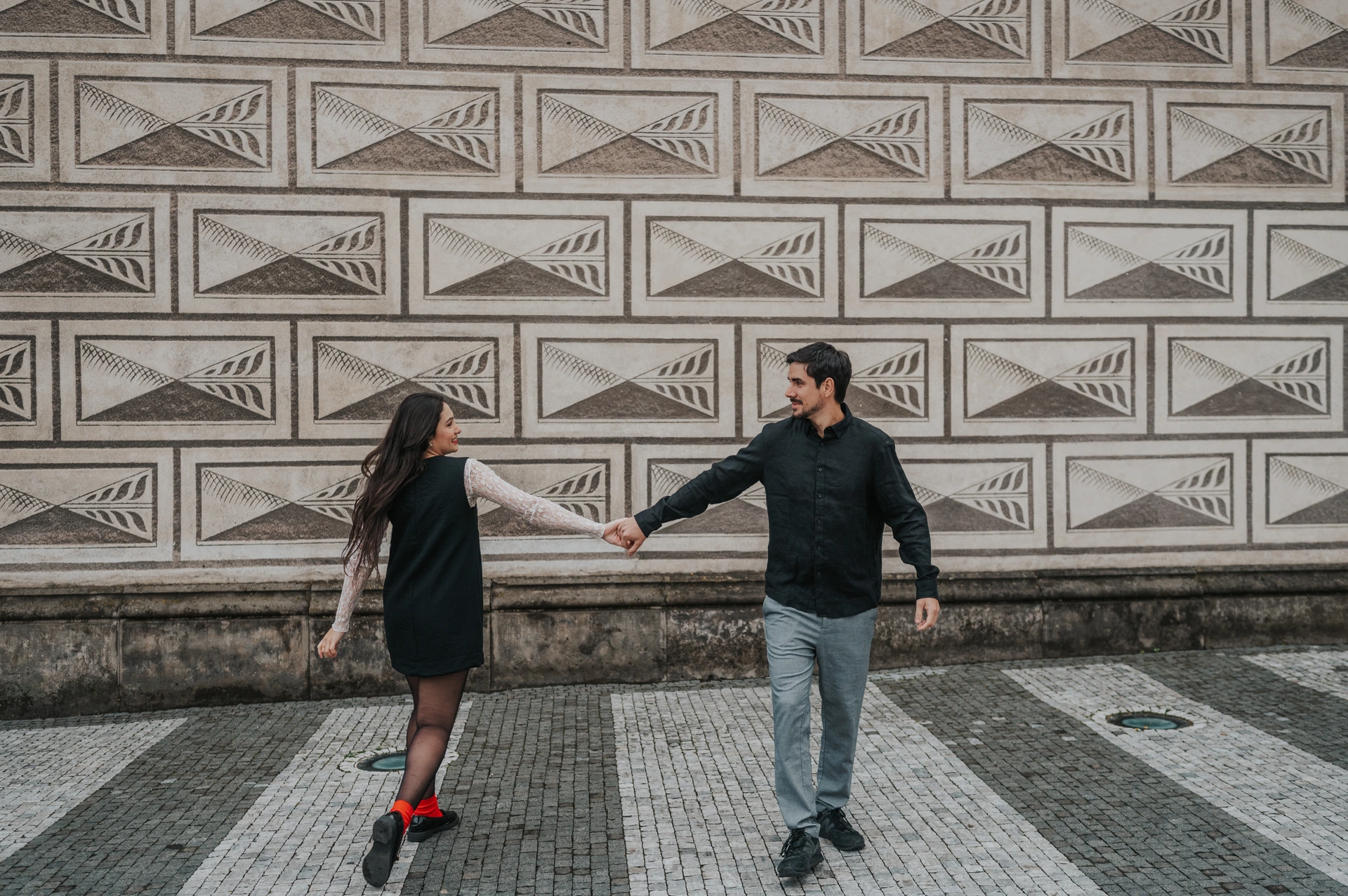 Prague engagement photoshoot with a playful hand-in-hand walk Couple reaching towards each other while walking hand in hand in front of a patterned wall during their engagement photoshoot in Prague.