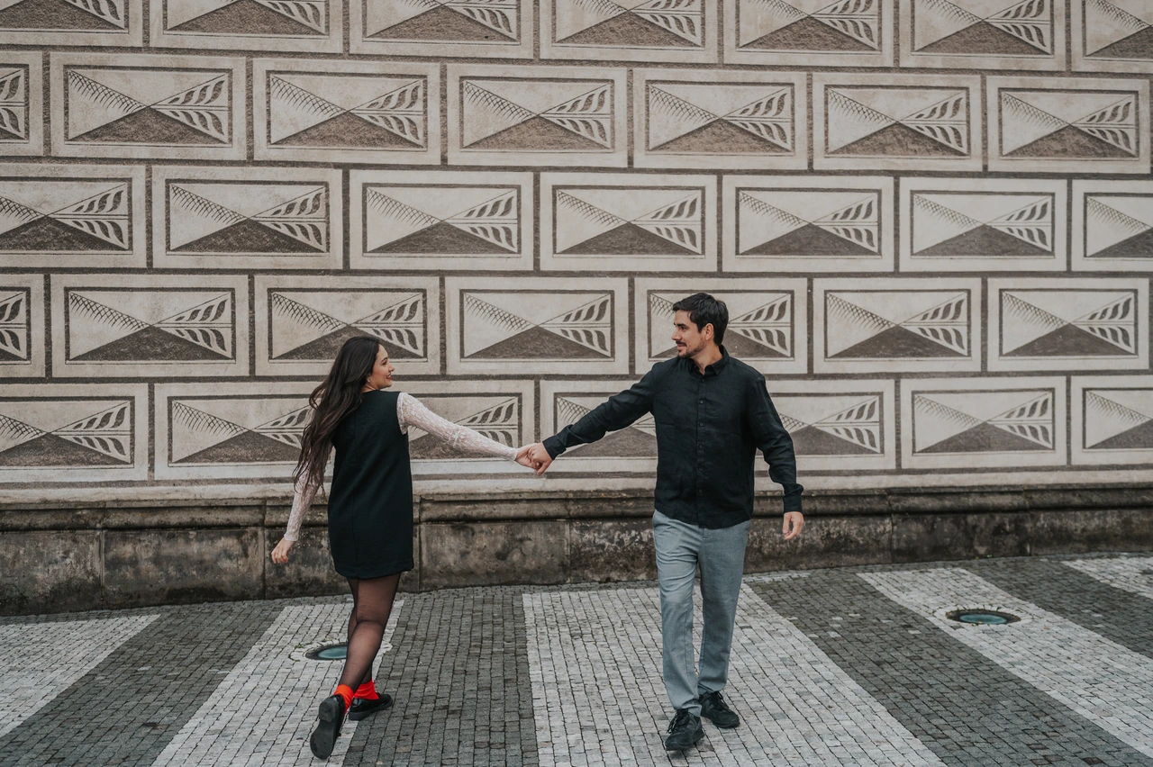Prague engagement photoshoot with a playful hand-in-hand walk Couple reaching towards each other while walking hand in hand in front of a patterned wall during their engagement photoshoot in Prague.