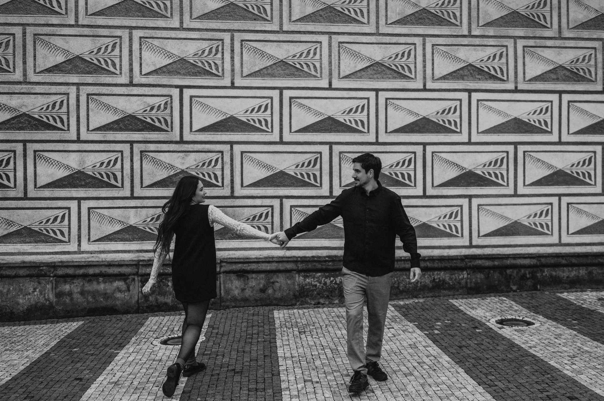 Black-and-white wide photograph of the couple walking hand in hand beside a patterned facade during their Prague engagement photoshoot.