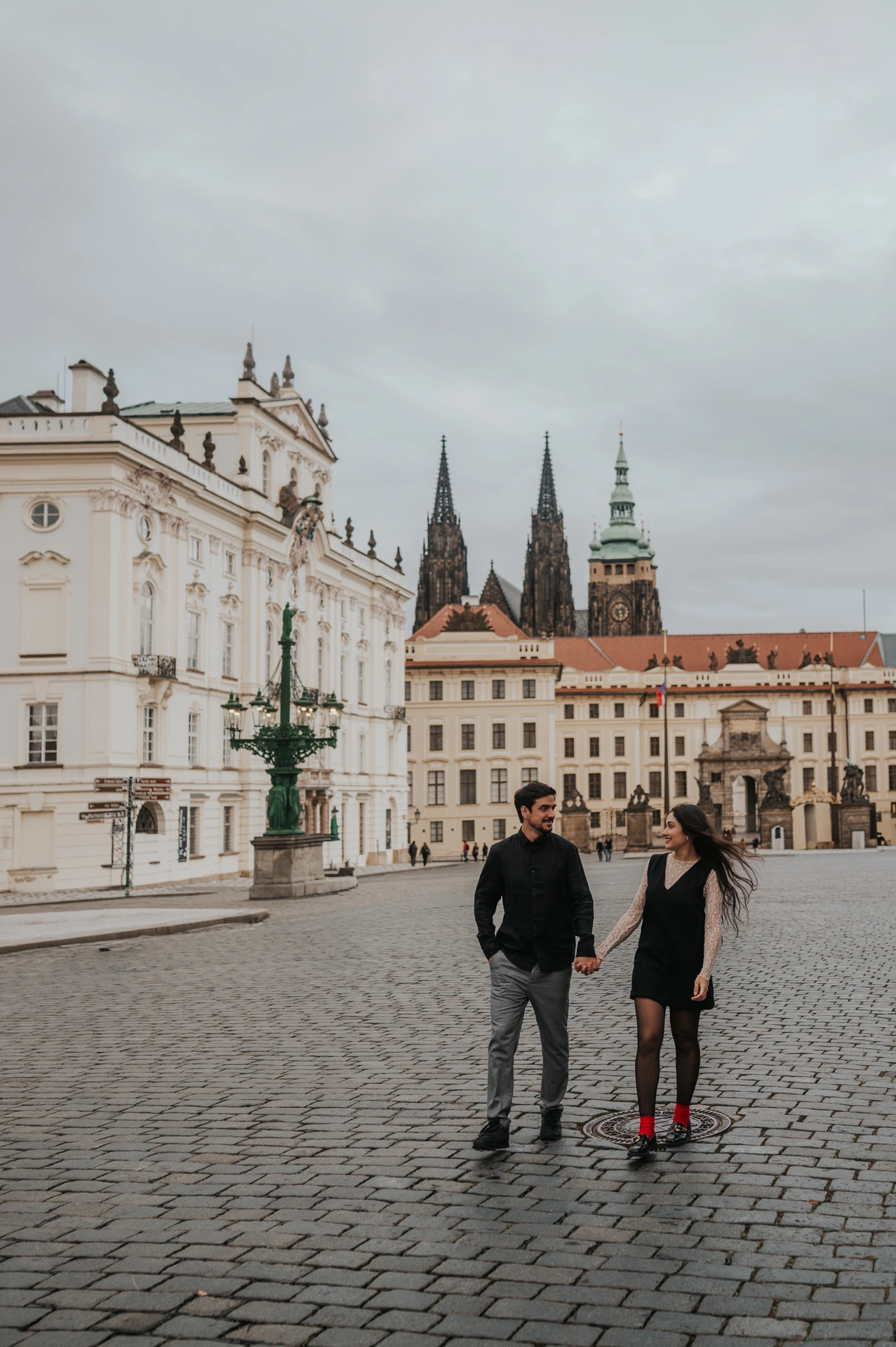 Couple walking hand in hand across the square, with Prague Castle in the background, during their engagement photoshoot.
