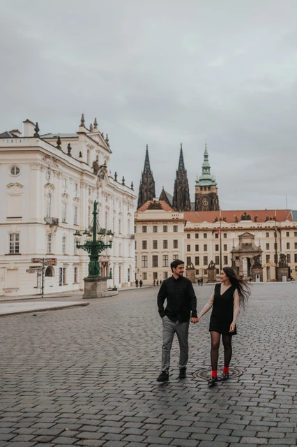Prague engagement photoshoot with the castle beyond the square Couple walking hand in hand across the square, with Prague Castle in the background, during their engagement photoshoot.