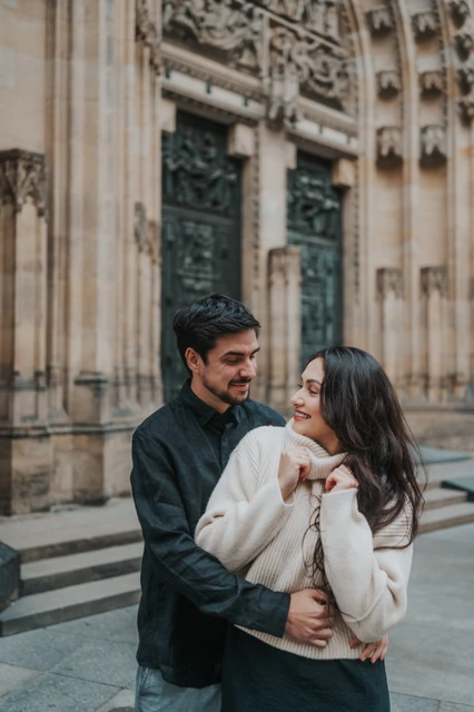 Prague engagement photoshoot with a relaxed cathedral embrace Couple smiling at each other in a close embrace in front of the cathedral, with the woman wearing cream knitwear, during their Prague engagement photoshoot.