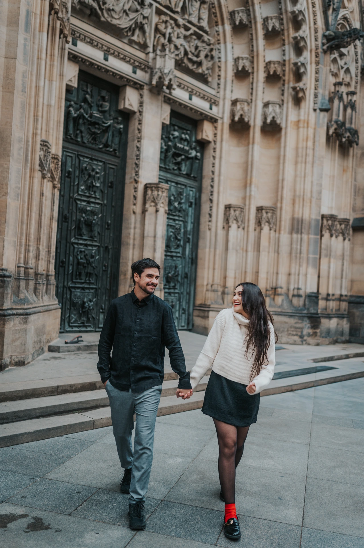 Couple walking hand in hand past the cathedral entrance, with the woman wearing cream knitwear, during their Prague engagement photoshoot.
