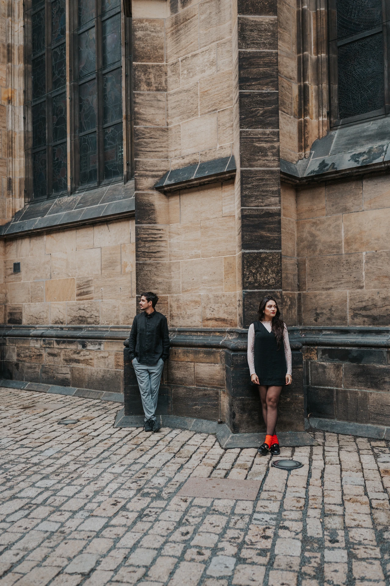 Couple standing apart against the sandstone cathedral wall below a tall window during their engagement photoshoot in Prague.